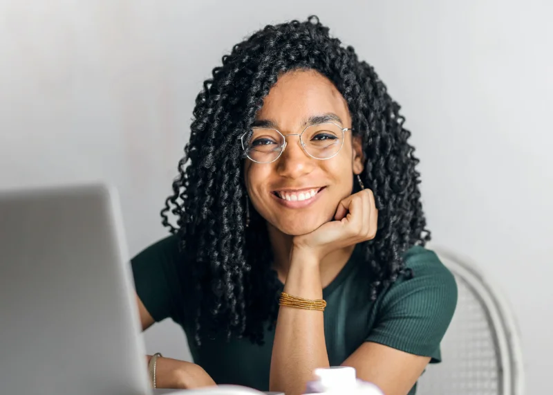 Jovem mulher de óculos em frente ao notebook sorri para a foto.