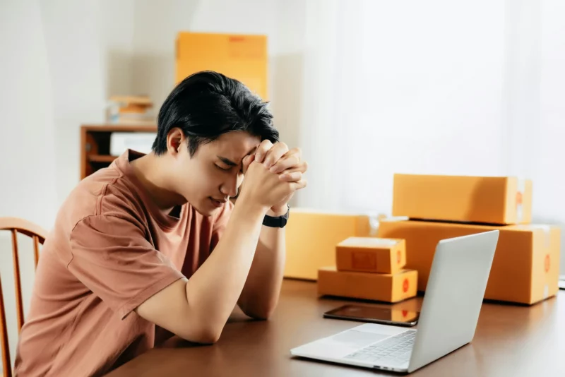 Homem em frente ao notebook com expressão preocupada e ao lado de algumas encomendas.